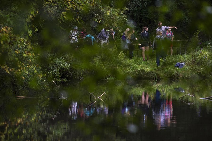 Students and a faculty member walk through a forest with a small body of water.