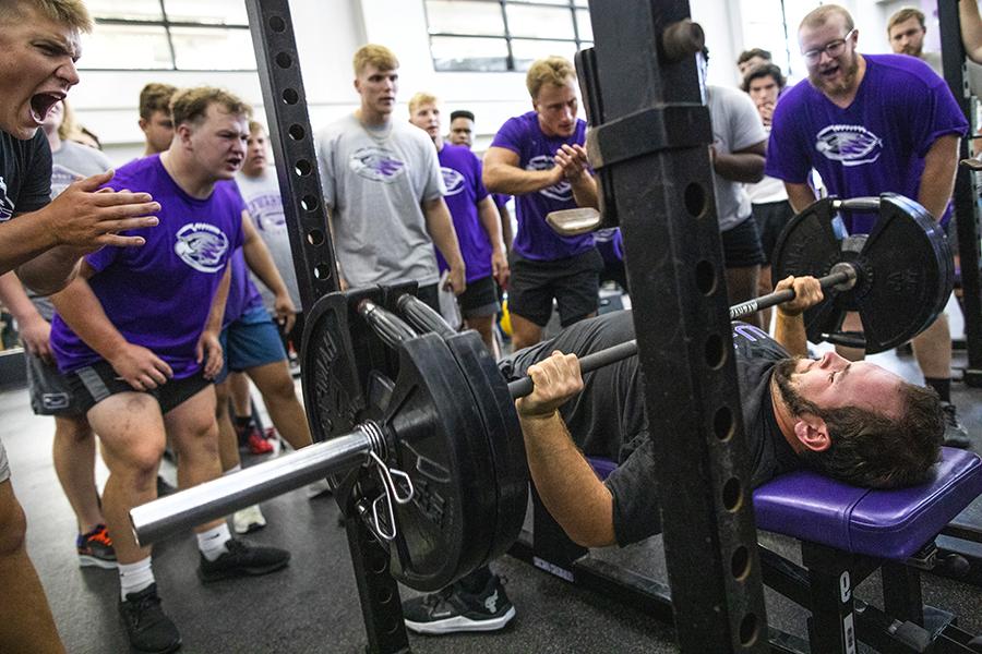 A group of crowd around another student benching a large amount of weight. 